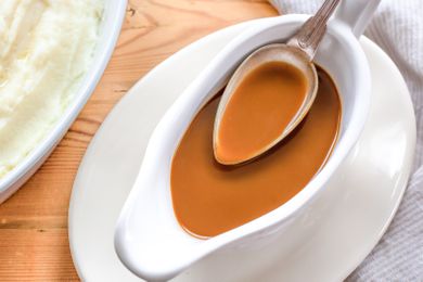 Overhead view of a gravy boat of gravy and a spoon full of gravy next to a plate of mashed potatoes and a white napkin all on a wooden tabletop