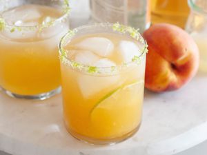 Two summery peach margaritas on a white tray with a whole peach next to it.