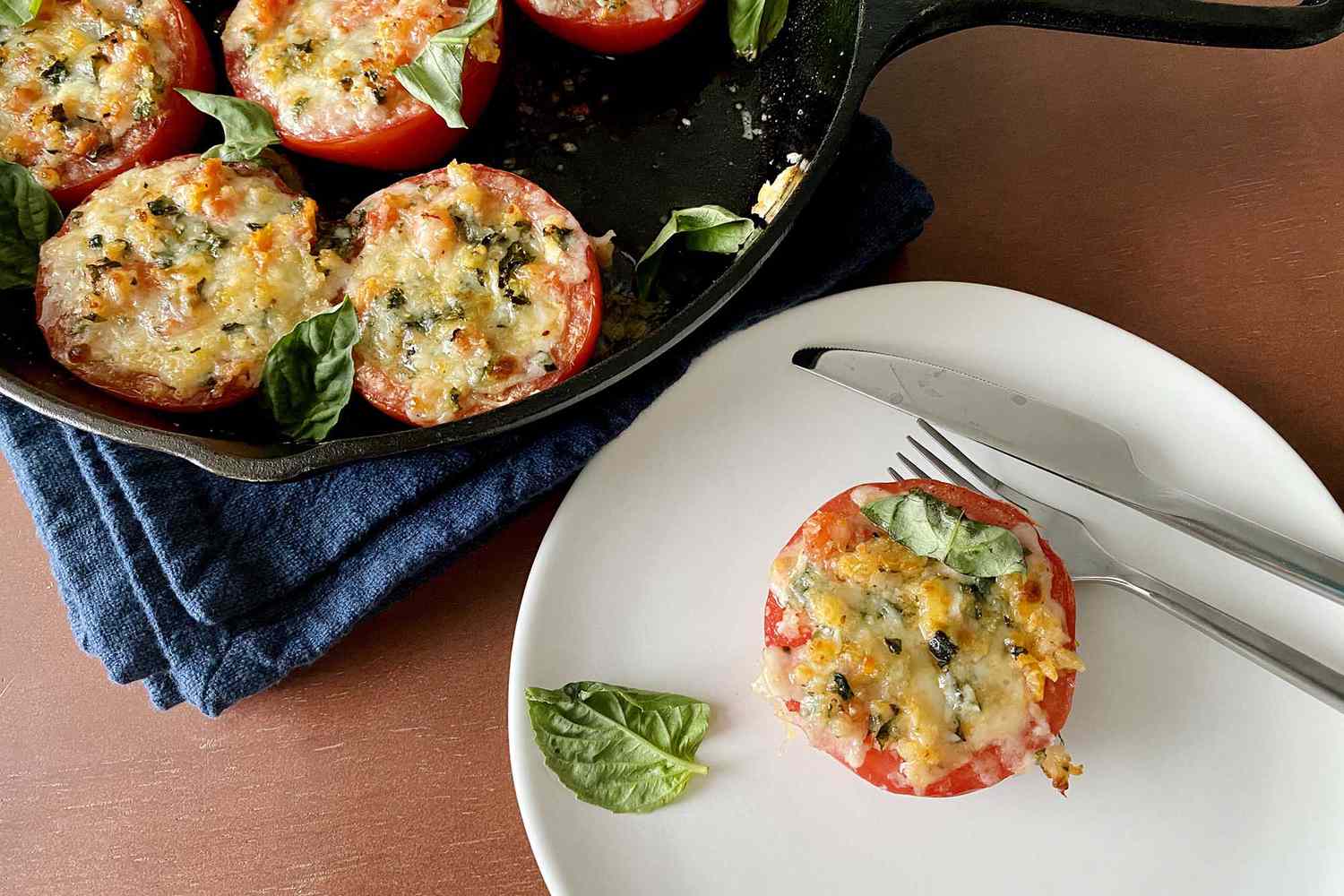 Easy baked stuffed tomatoes on a plate with a knife and fork. A cast iron skillet with additional stuffed tomatoes are to the left.