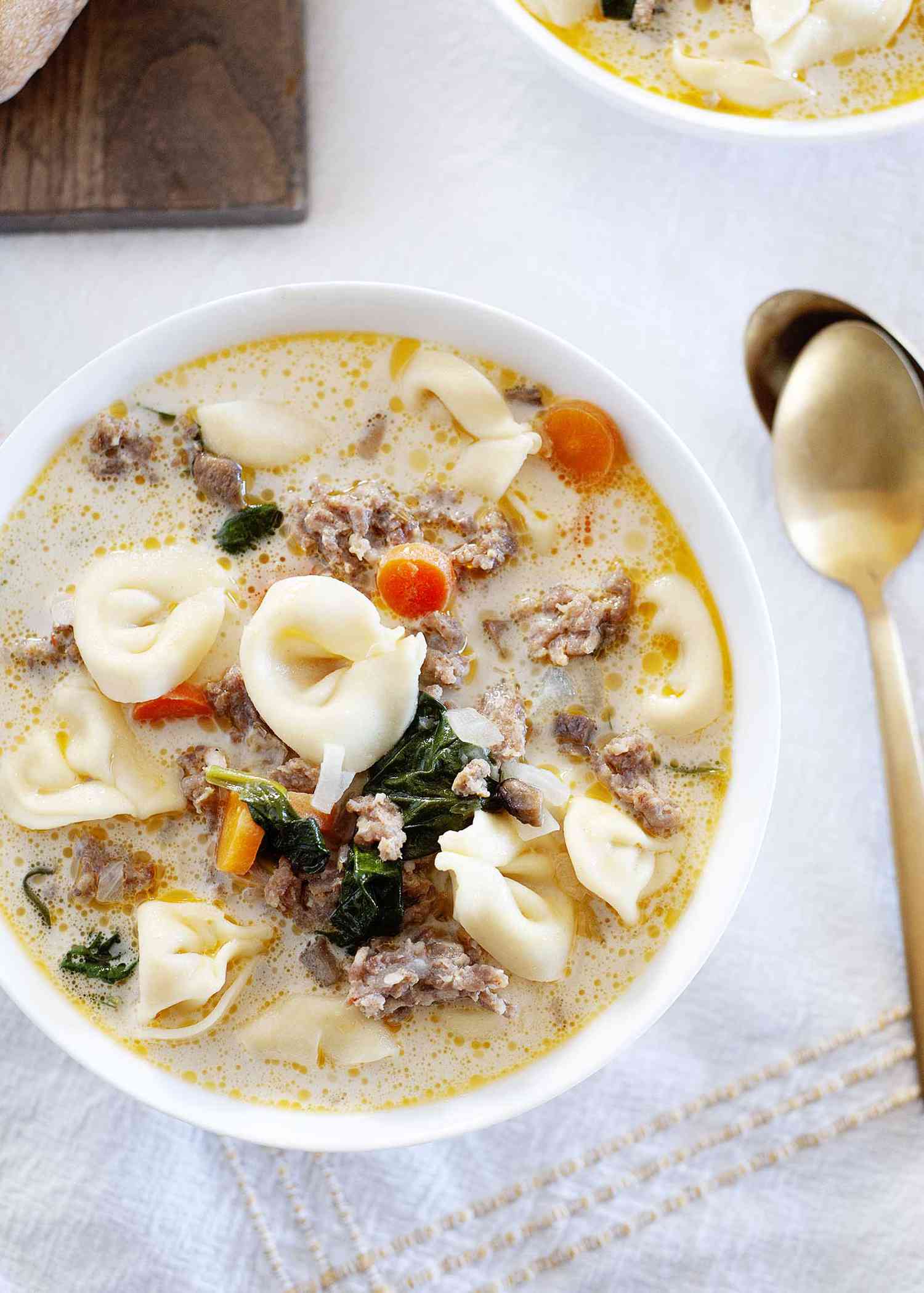 Creamy sausage tortellini soup in a white bowl with sausage, tortellini and carrots visible in the bowl. The table is covered in a white tablecloth and next to the bowl are two gold spoons. A second bowl of soup and a wooden cutting board are in partial view in the background.