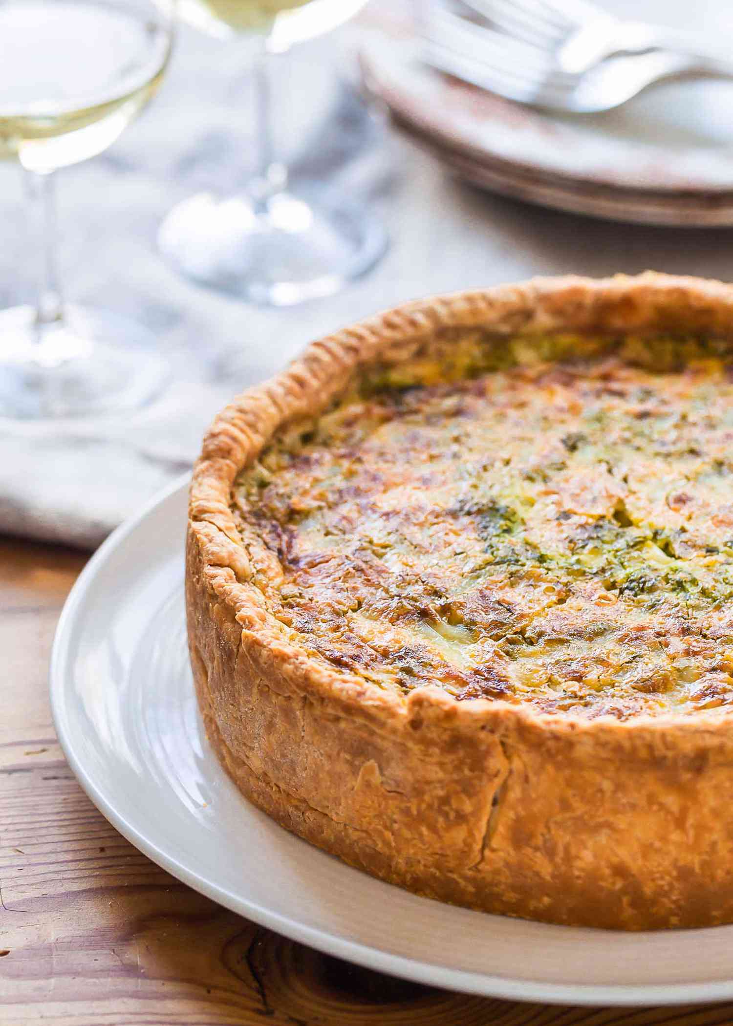 A white platter is on a white plate set on a wooden table. The platter has a homemade deep-dish bacon and cheddar quiche on it. The crust and filling are golden brown and some parsley is visible in the filling. Two wine glasses, plates and forks are above the quiche on a cream colored table linen.