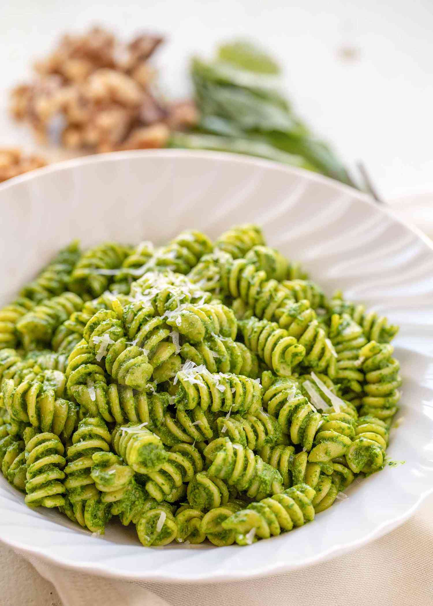 A white bowl with rotini and kale pesto inside. Walnuts and basil are in the background.