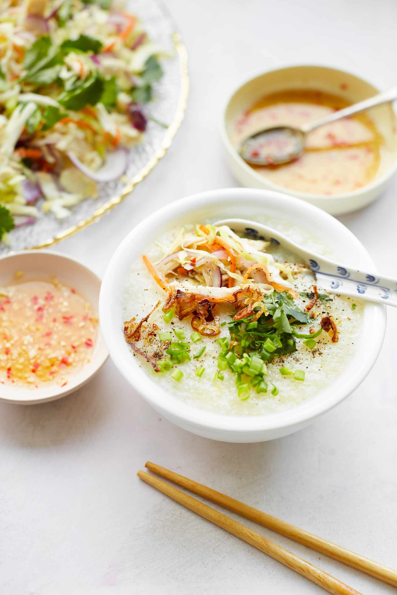Bowl of Chào Gá (Vietnamese Rice Porridge) with Vegetables and Herbs, Surrounded by Platter of Vegetables and Bowls of Sauce
