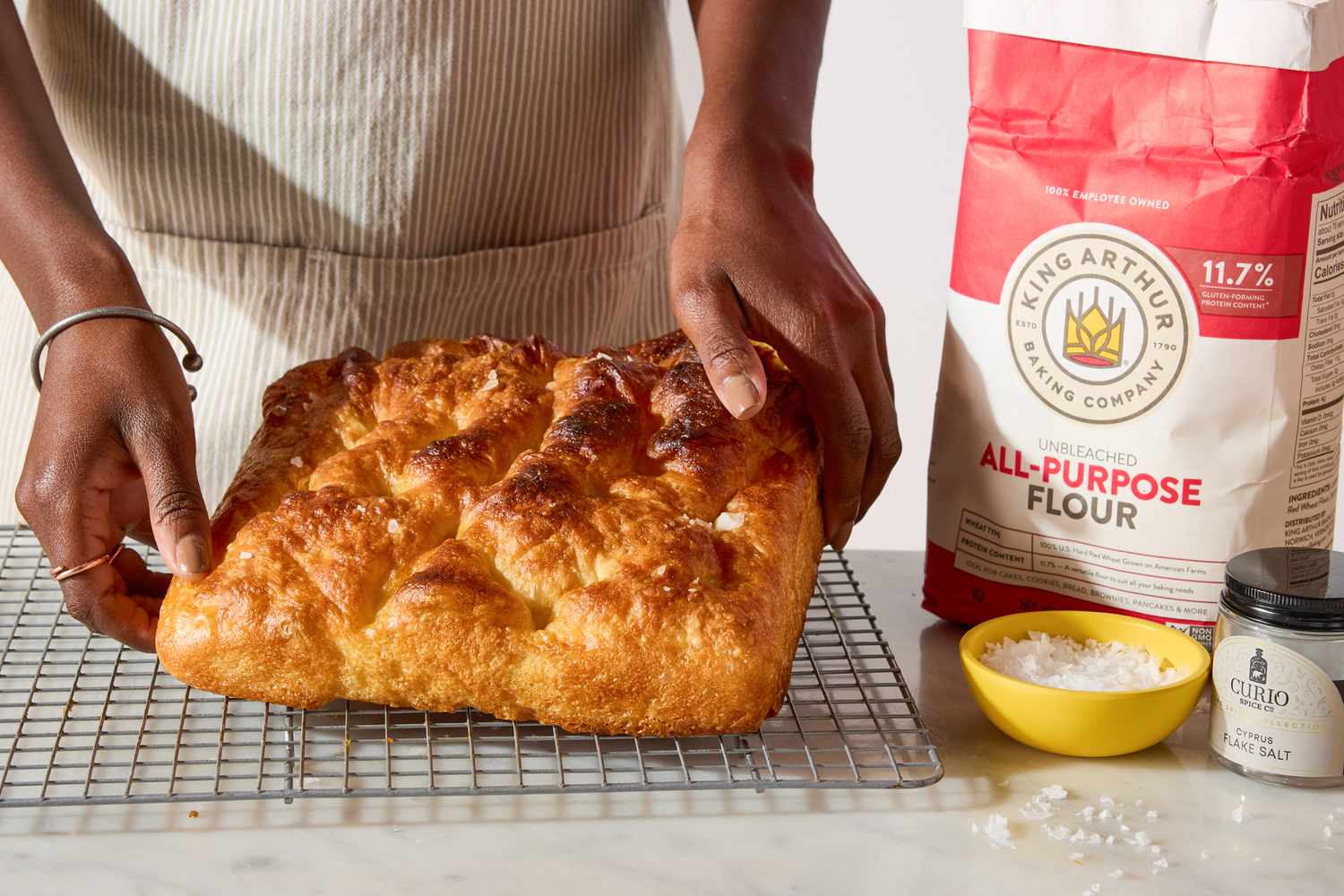 hands placing King Arthur Flour’s Big and Bubbly Focaccia on a cooling rack, flour and salt on the side