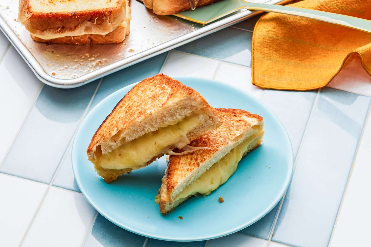 Sheet Pan Grilled Cheese Sandwich Cut in Half and on a Light Blue Plate Next to a Table Napkin and a Sheet Pan With More Grilled Cheese Sandwiches