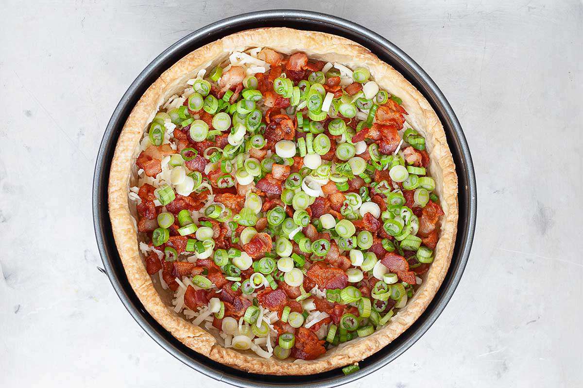 A springform pan with a golden brown crust has chopped scallions and crispy chopped bacon at the bottom of it.