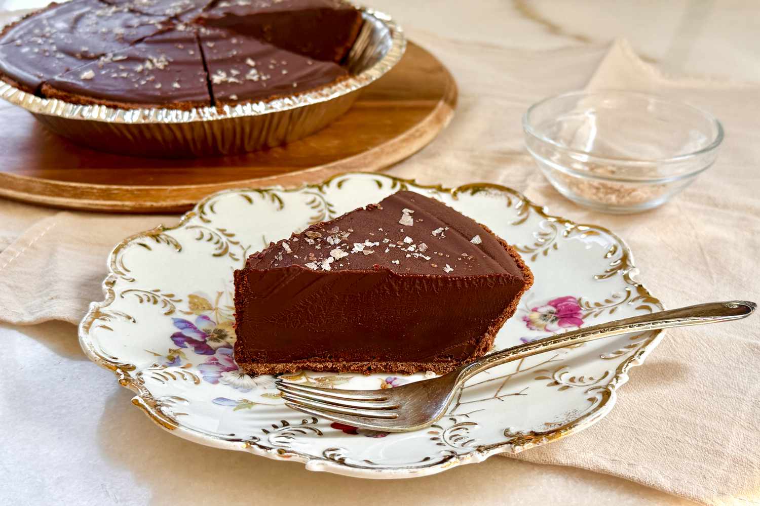 A slice of chocolate tart on a decorative plate with a fork, tart in the background