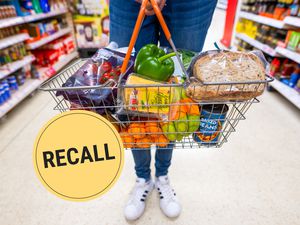 A person from the waist down wearing jeans and Adidas sneakers standing in the middle of a grocery store aisle holding a full shopping basket. A "Recall" badge is superimposed over the photo.