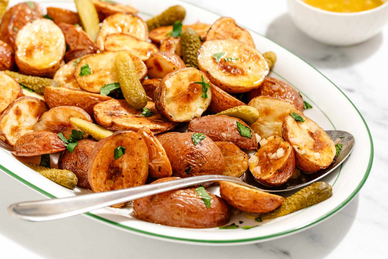 Closeup view of a serving spoon on an oval platter of roasted potato halves and pickles topped with parsley