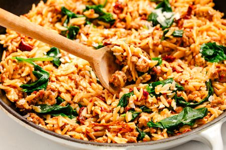 Angled view of a wooden spoon in a skillet of orzo, italian sausage, sun-dried tomatoes and baby spinach