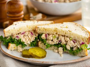 tuna salad sandwich (cut in half) on a plate with a few pickles, and in the background, more tuna salad in a bowl