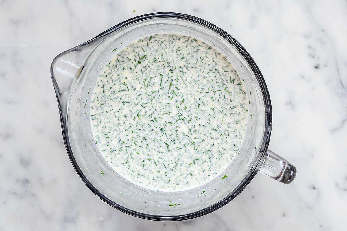 A large glass liquid measuring cup is filled halfway with a creamy filling with chopped parsley inside.
