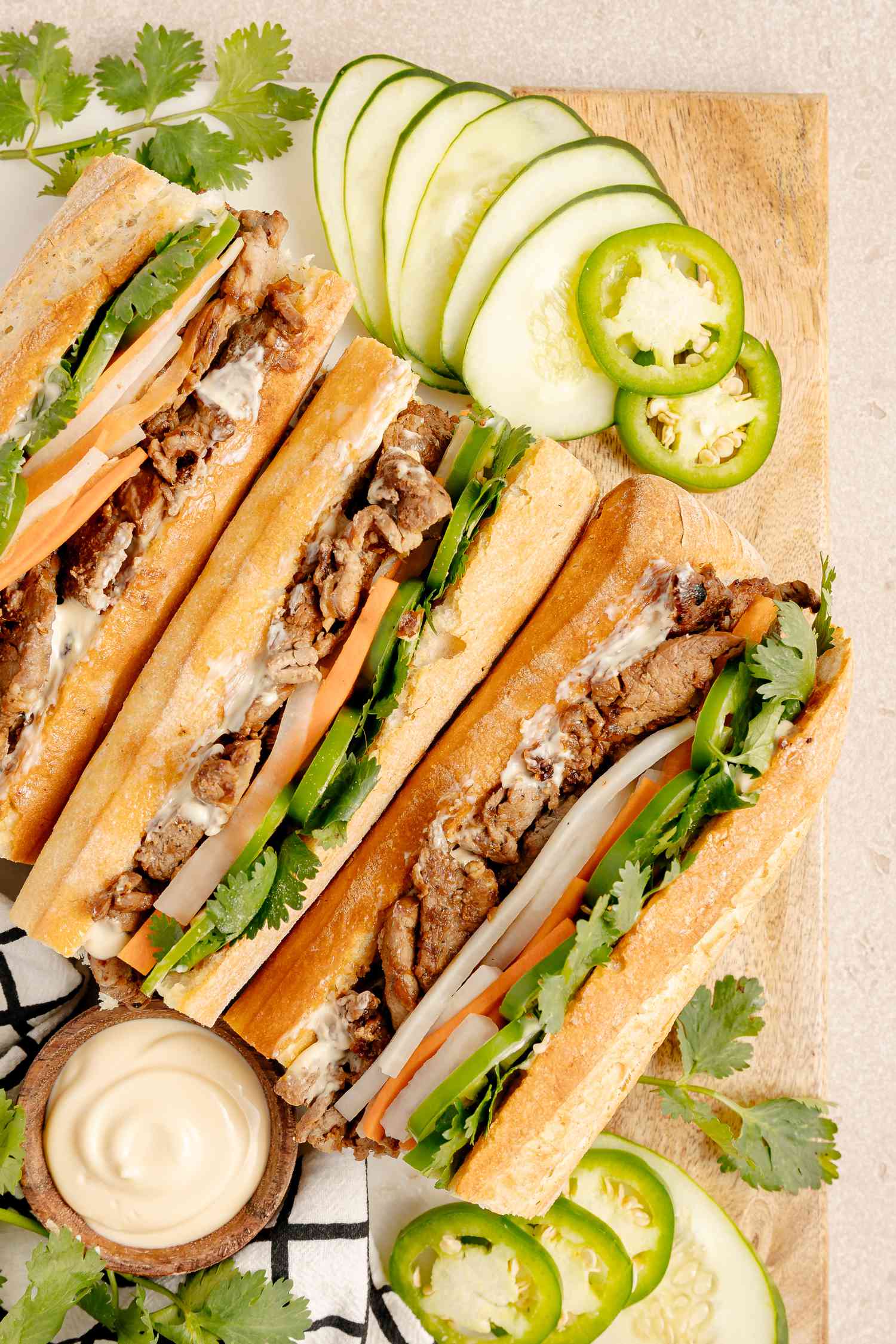 Pork Banh Mi Surrounded by Slices of Cucumber, Jalapenos, Cilantro, and a Bowl of Mayo