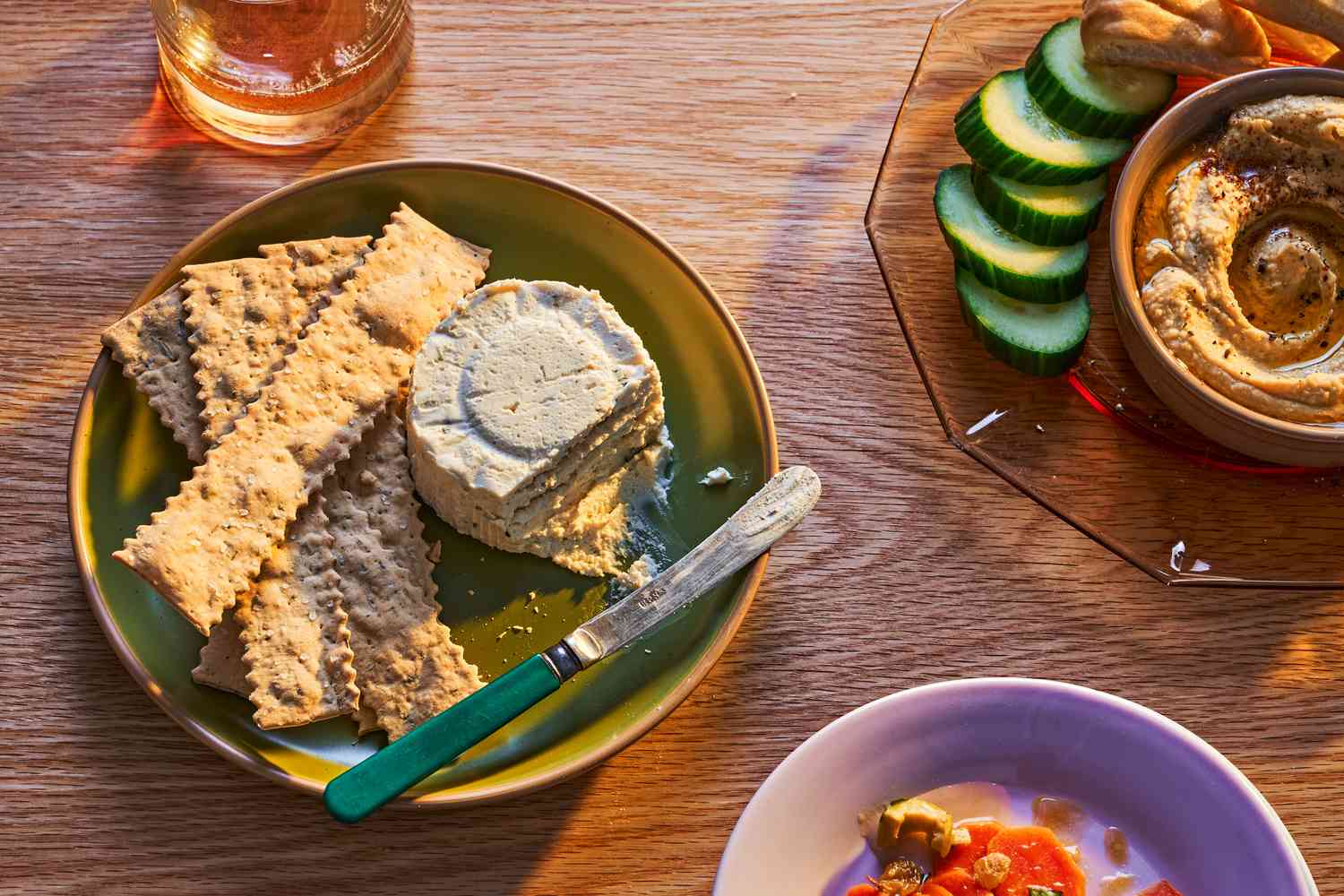 A plate with Boursin cheese and crackers, surrounded by a glass of wine, hummus and cucumbers
