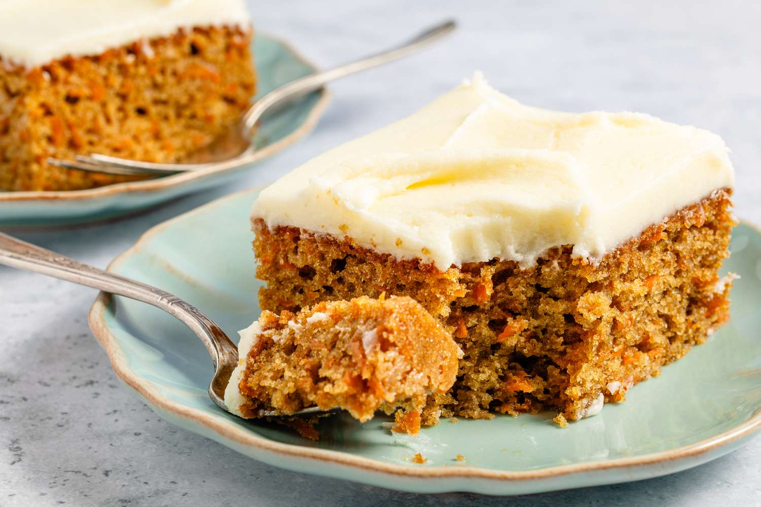 Slice of carrot cake with cream cheese frosting on a plate, fork holding a small bite of cake.