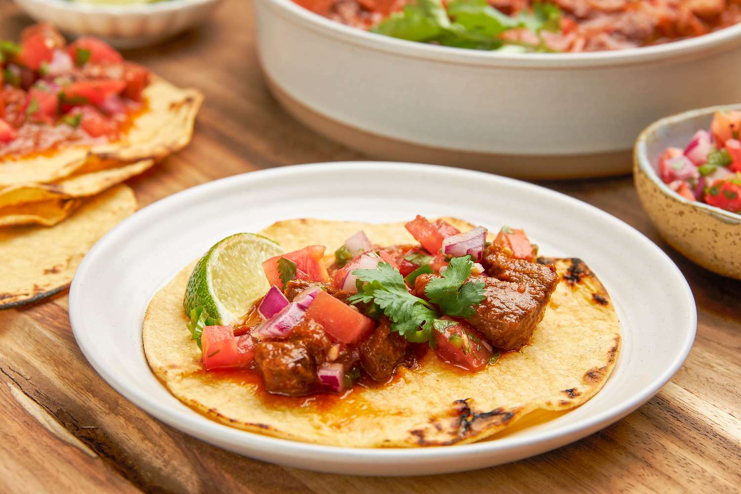 A toasted tortilla with carne picada, pico de gallo, and a lime wedge on a plate, and in the surroundings, bowls of toppings (carne picada and pico de gallo) and tortillas on the wooden board