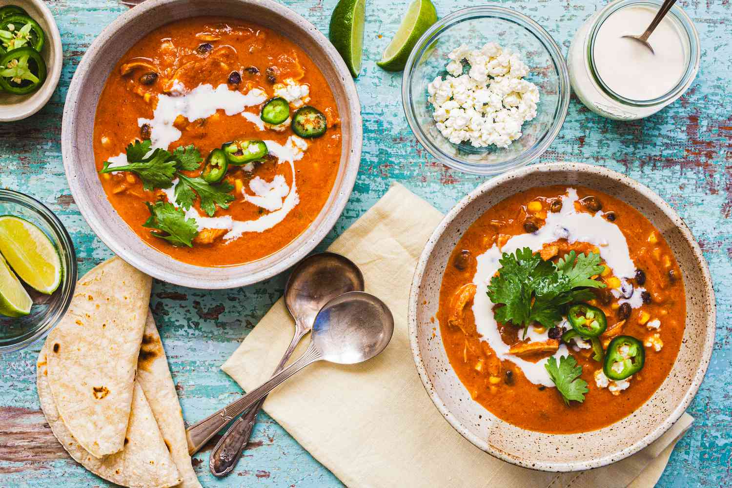 Two Bowls of Creamy Chicken Enchilada Soup Garnished with Cilantro, Crema, and Jalapenos at a Table Setting with Two Spoons on a Table Napkin, Bowls of Toppings (Jalapenos, Lime Wedges, Cotija, and Crema), and Tortillas