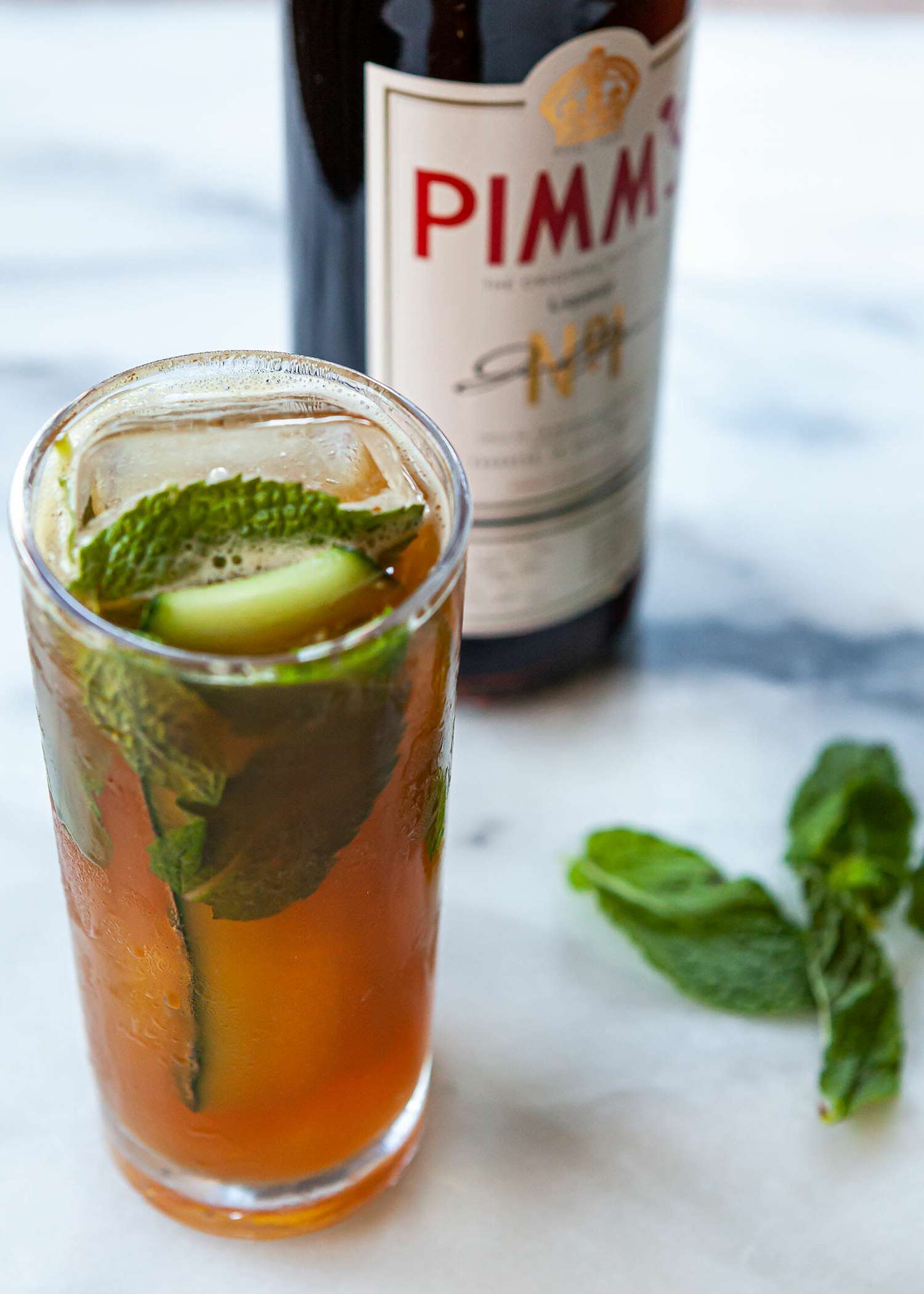 A tall glass with a Pimms cup cocktail is on a counter with mint leaves and a bottle of Pimms no 1 behind it.