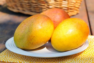 Three ripe mangos arranged on a white plate with a wicker basket in the background