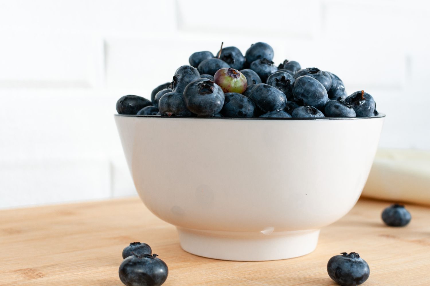 Side view of a white bowl full of blueberries and several blueberries scattered around it all on a bamboo countertop against a white tile background