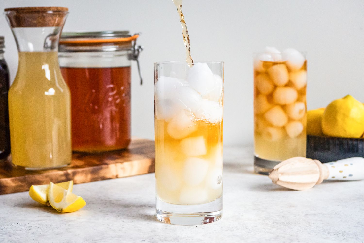 Assam Tea Poured Into Glass Filled With Ice and Some Lemonade. In the Background, Another Glass With Tea, Ice, and Lemonade, a Bowl of Lemons, and a Citrus Reamer, and on the Left, Two Jars (One With Lemon Juice and Another With Black Tea) on a Wooden Board and Two Lemon Wedges for Arnold Palmer Recipe
