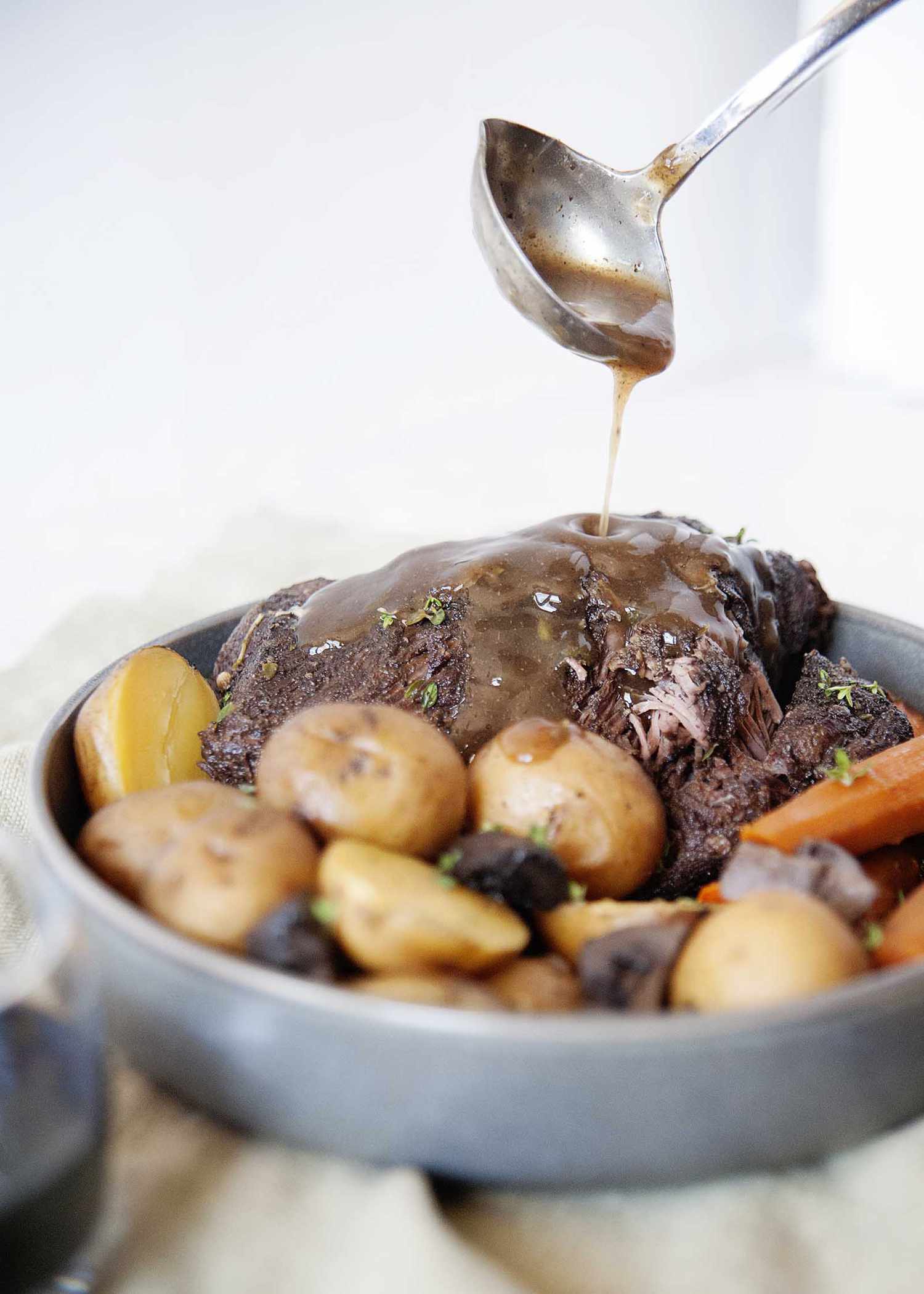Ladling gravy over a platter full of pot roast, potatoes and carrots. 