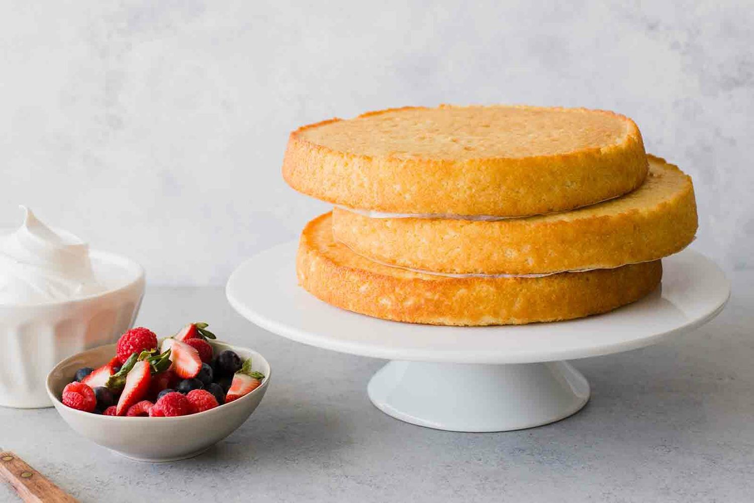 A stack of cake layers on a cake stand with berries and whipped cream on the side