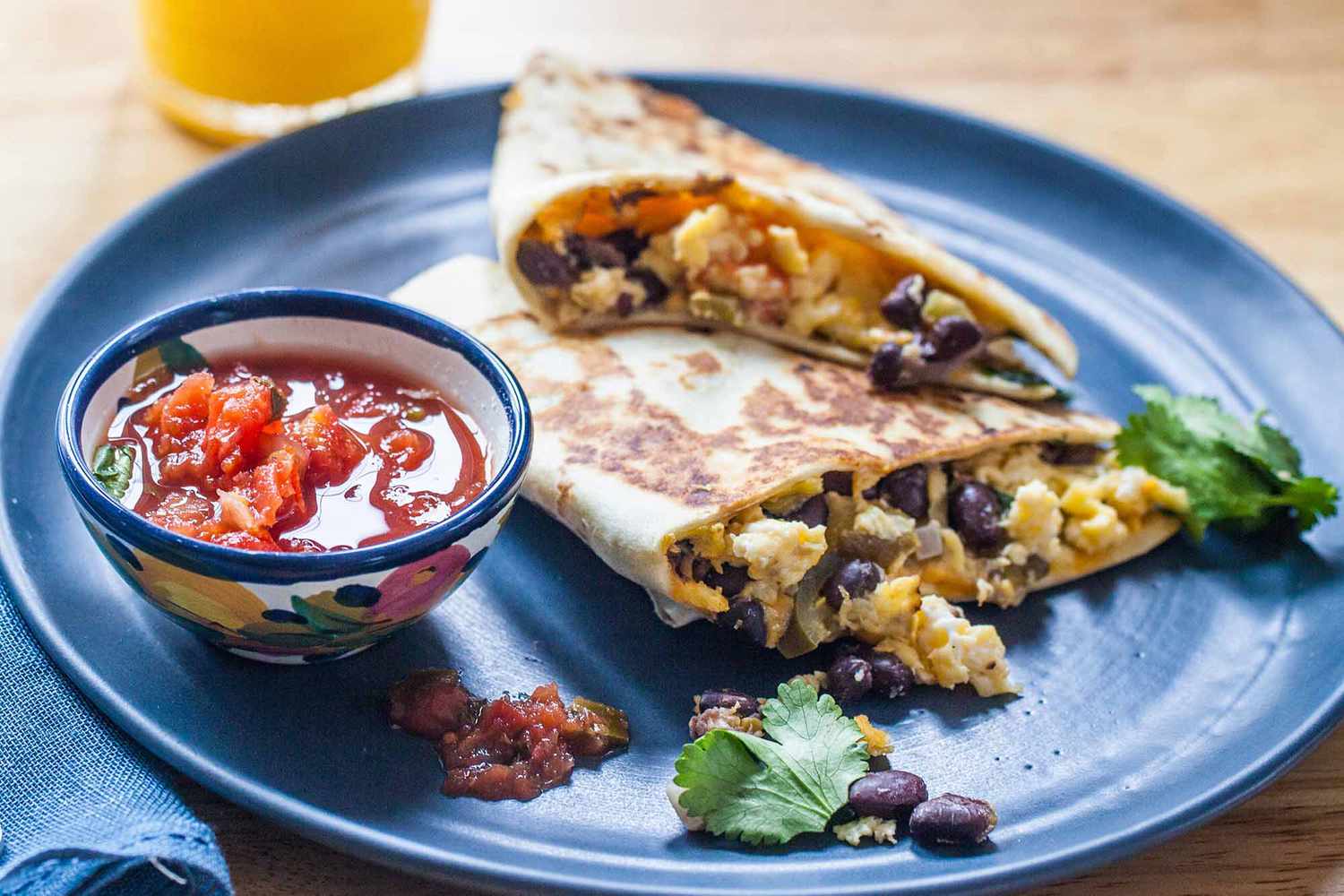 Side view of two simple breakfast quesadillas on a blue plate with a bowl of salsa to the left and a glass of orange juice above. The scrambled eggs, black beans and cilantro are visible inside the quesadilla.