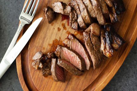 Overhead view of sliced steak cooked in the oven and silverware on a wooden cutting board.