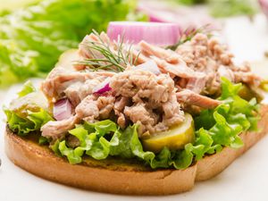 Open-faced tuna salad sandwich on toast with lettuce, pickles, and dill, garnished with red onion slices