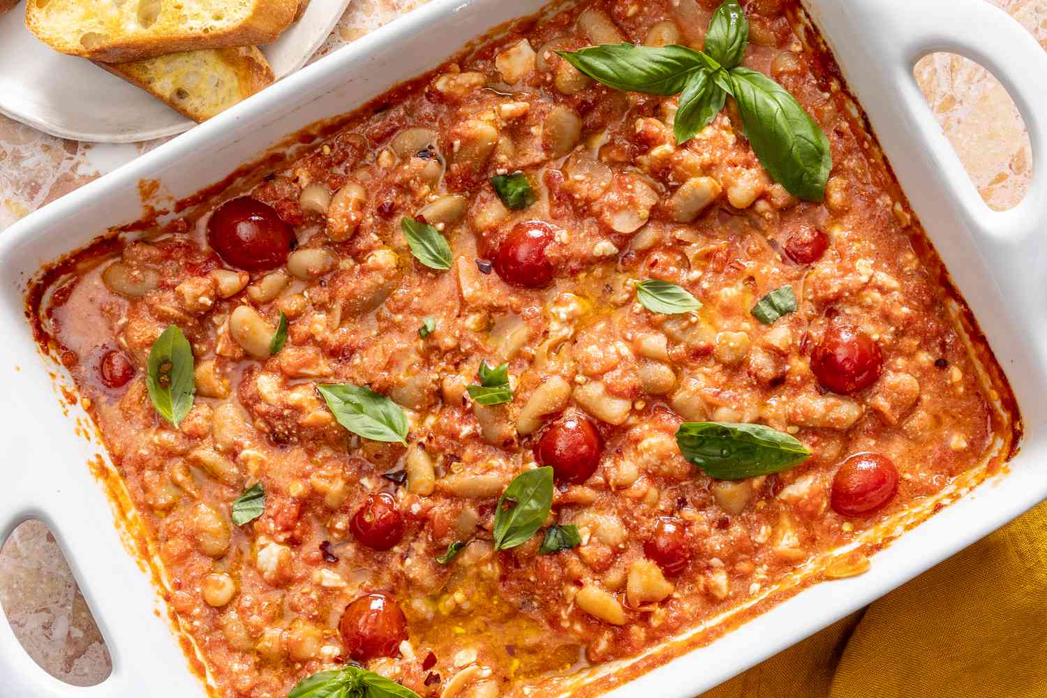 Overhead view of a white baking dish with a garlic, tomato, cannellini bean and feta mixture next to a plate of french bread slices 