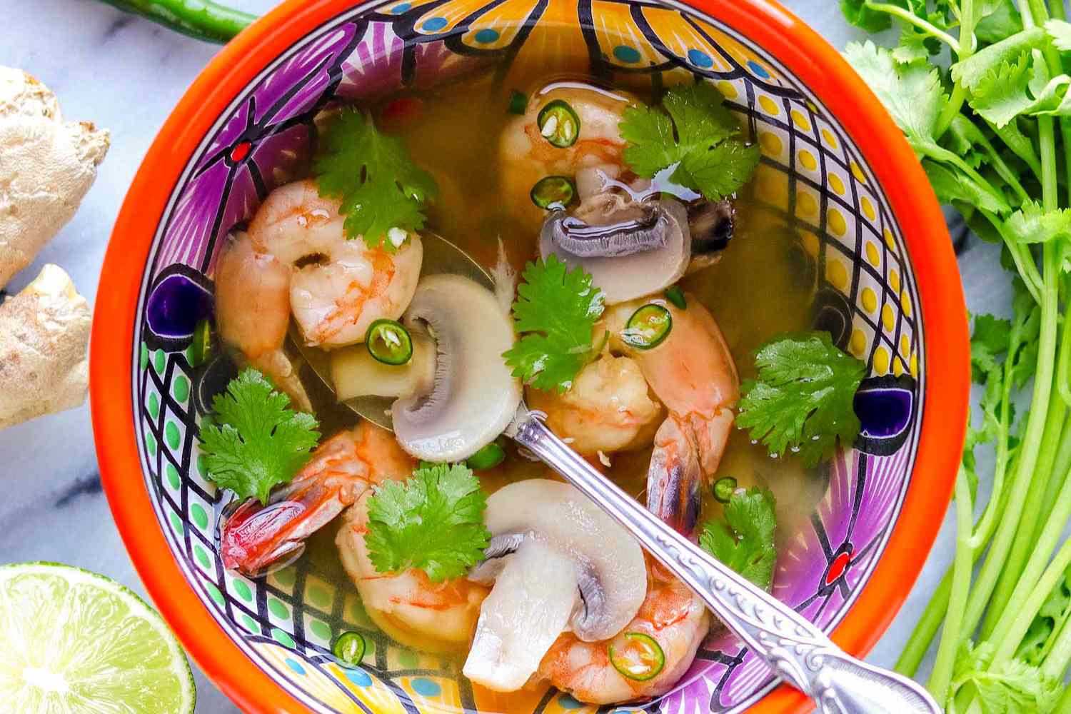 A bowl of shrimp tom yum with cilantro, halved lime, and ginger nearby