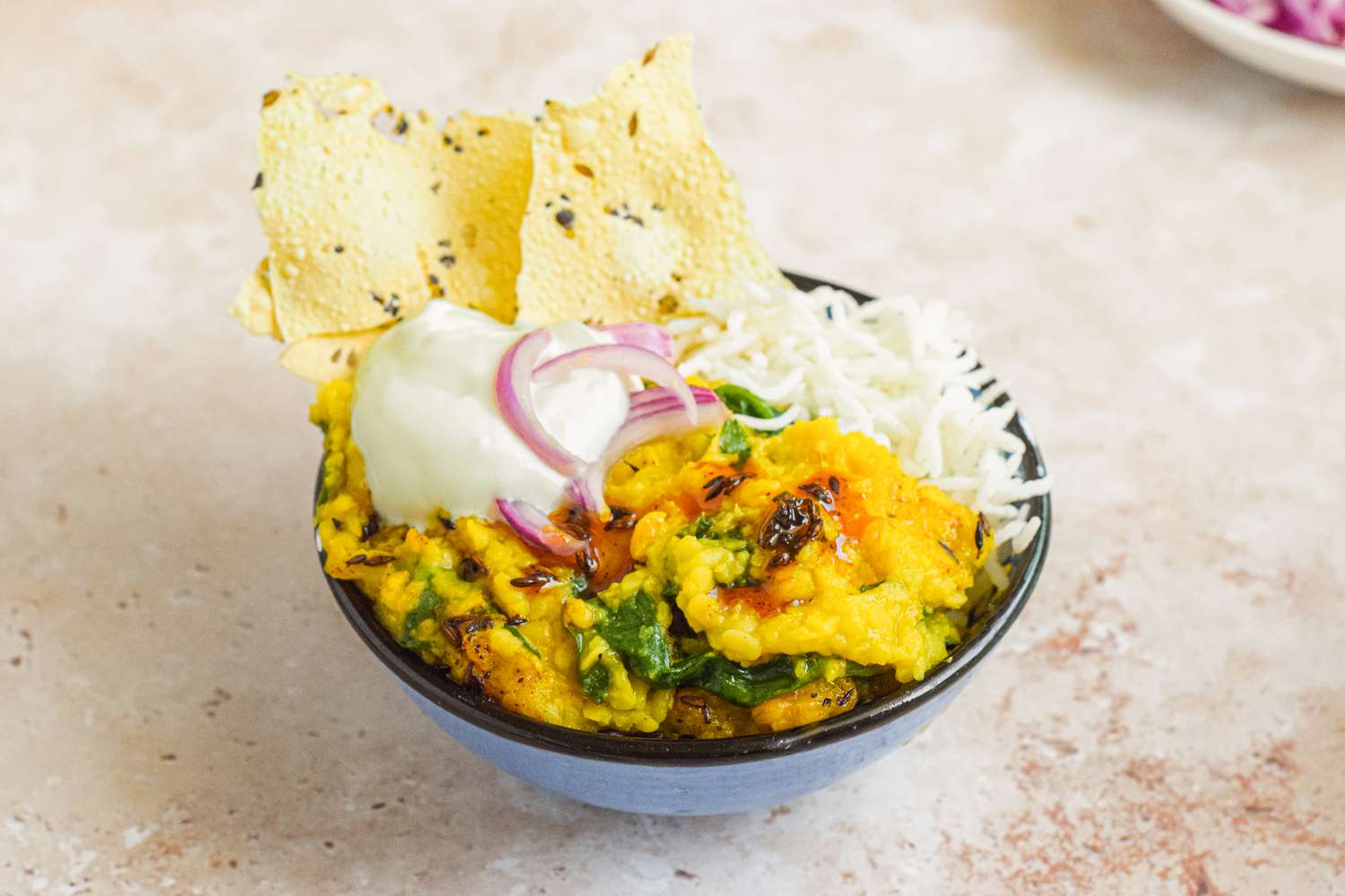 Bowl of Daal Palak over Rice Topped with Onions and Yogurt, Served with Pieces of Papad