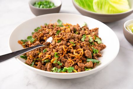 Plate of ground beef larb with sliced herbs, garnished with fried onions, surrounded by small bowls of lettuce and lime wedges