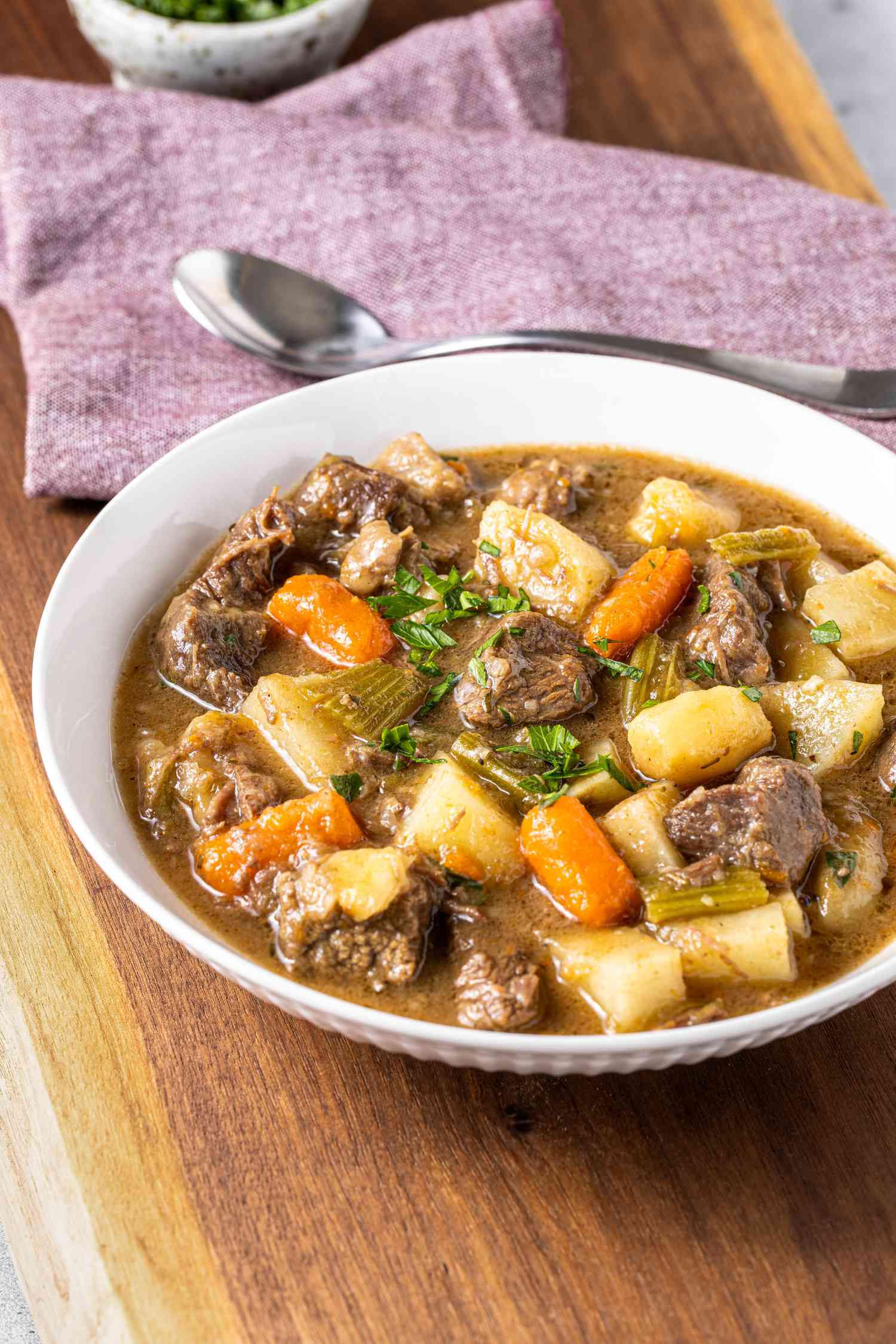 Bowl of Easy Beef Stew Toped with Some Parsley and in the Background, a Kitchen Towel with a Spoon on Top and a Small Bowl of Parsley