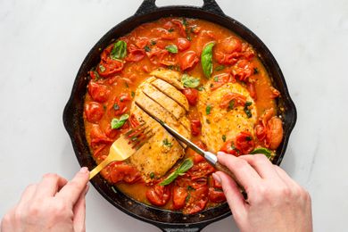hands with knife and fork slicing cooked chicken breasts in a tomatoey sauce in a cast iron skillet