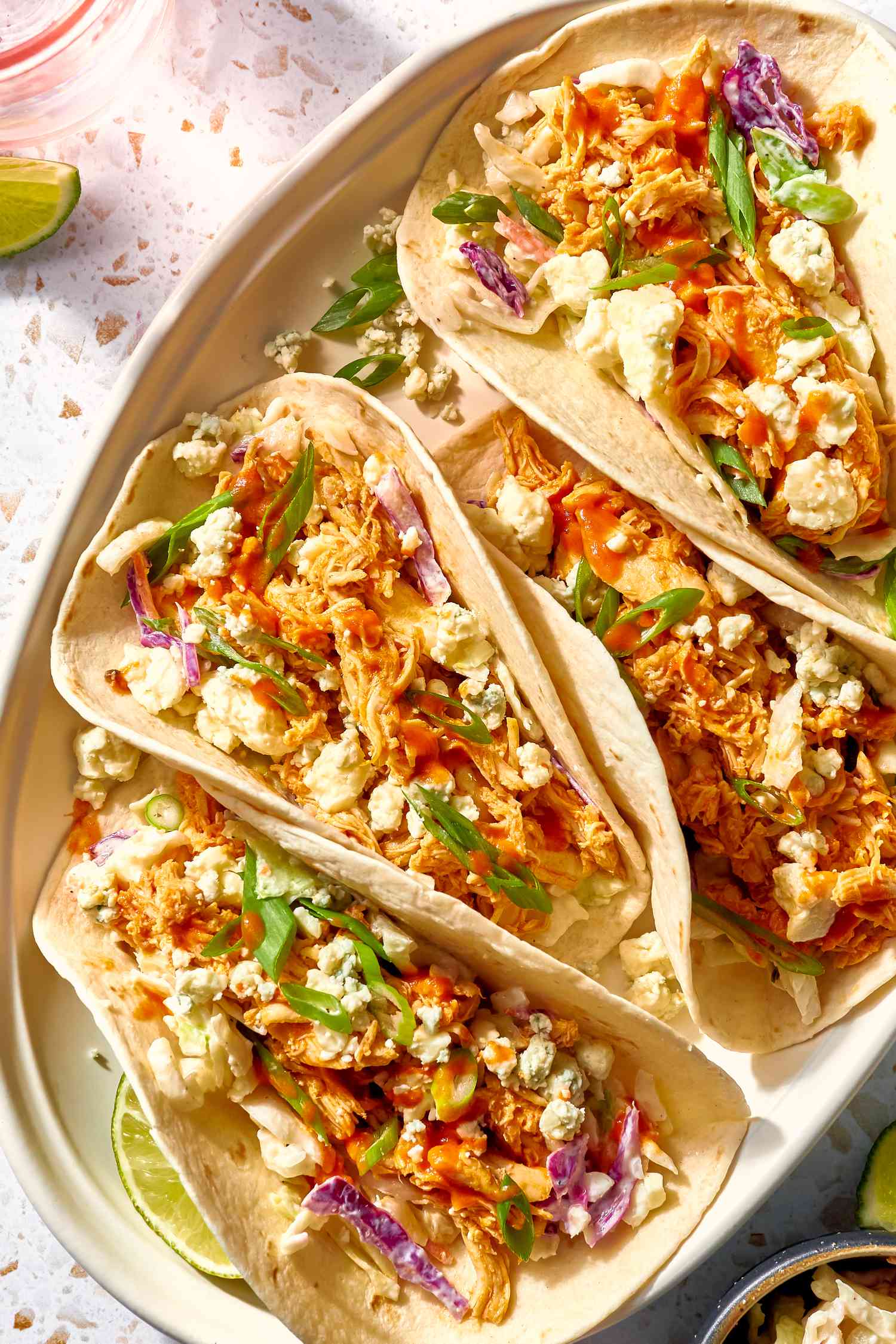 Buffalo Chicken Tacos Topped With Blue Cheese Crumbles and Green Onion Slices and Next to a Lime Quarter, All on a Platter, and in the Surroundings, Another Lime Slice on the the Counter, a Small Bowl With More Cheese, and a Glass of Water