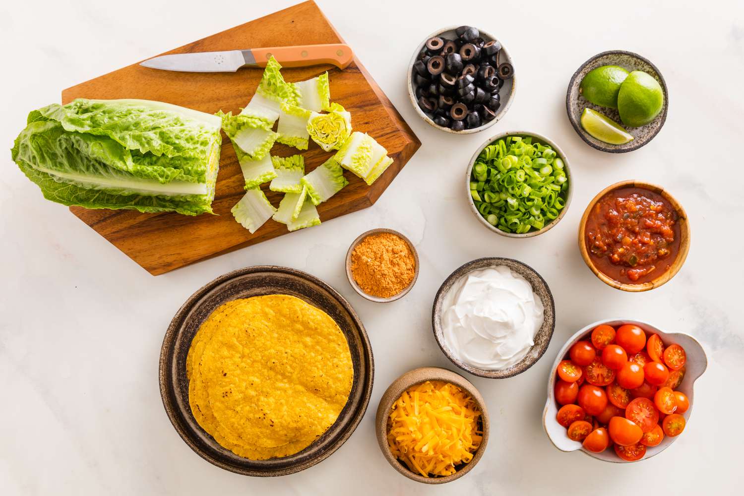 Taco Salad Ingredients Prepped and in Bowls. From L to R: One Head of Romaine on a Cutting Board With Some of It Cut Into Bite Size Pieces, a Plate With Tostadas, a Bowl of Sliced Olives, a Bowl With Sliced Green Onions, a Bowl of Shredded Cheese, a Bowl of Sour Cream, a Bowl of Salsa, and a Bowl of Halved Grape Tomatoes