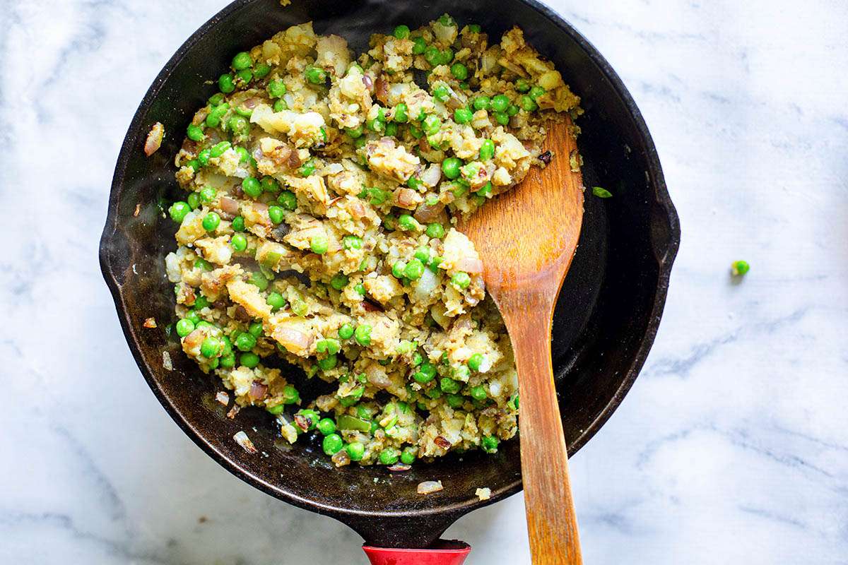 Onions, spices, peas, potatoes and jalapenos being toasted in a black skillet for vegetable samosa filling.