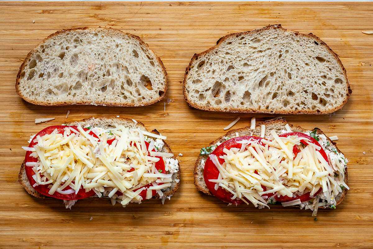 How to make a tuna melt. Four pieces of sturdy bread laid out on a wooden cutting board and two of the pieces have tuna salad, sliced tomatoes and shredded cheese on top.