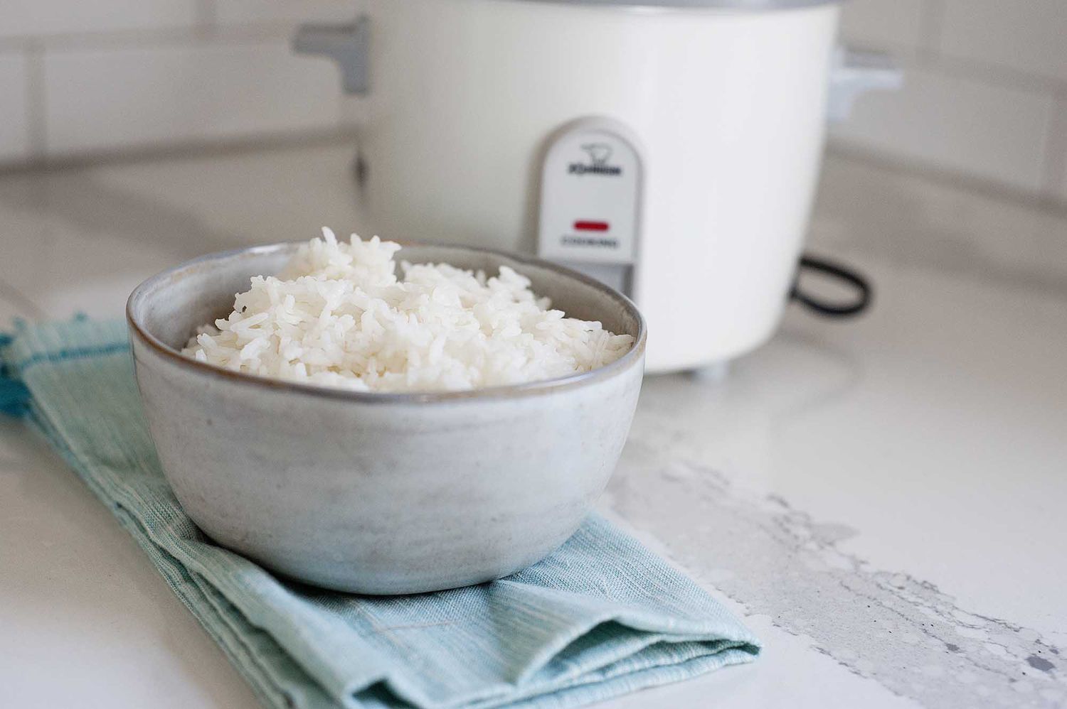 Horizontal image of a ceramic bowl filled with white rice on a white counter. A teal dish towel is under the bowl of rice and a white rice cooker is in partial view behind the bowl.