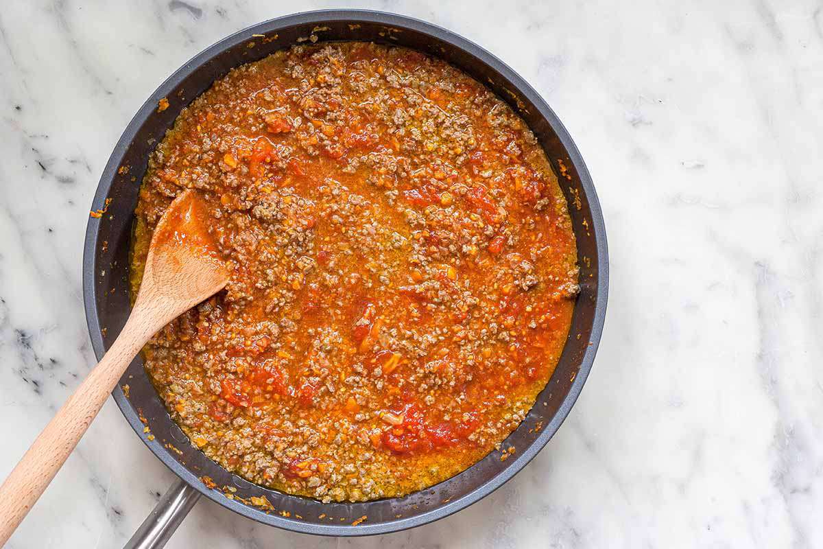 A homemade meaty bolognese sauce in a skillet on a marble countertop with a wooden spoon.