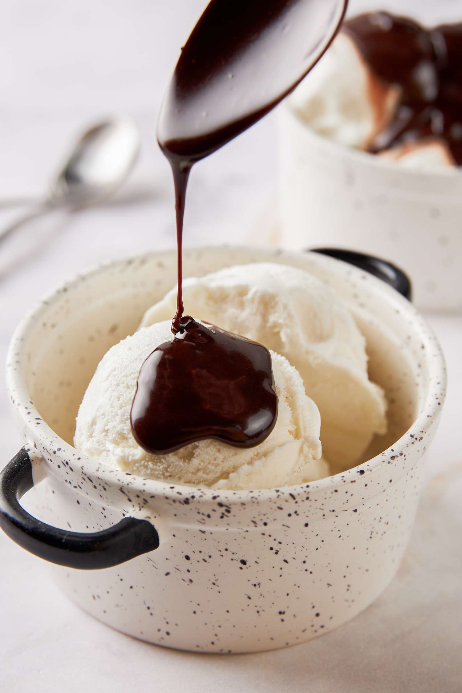 Spoonful of hot fudge sauce poured onto vanilla ice cream in a bowl with handles, and in the background, another bowl of ice cream with hot fugde sauce 