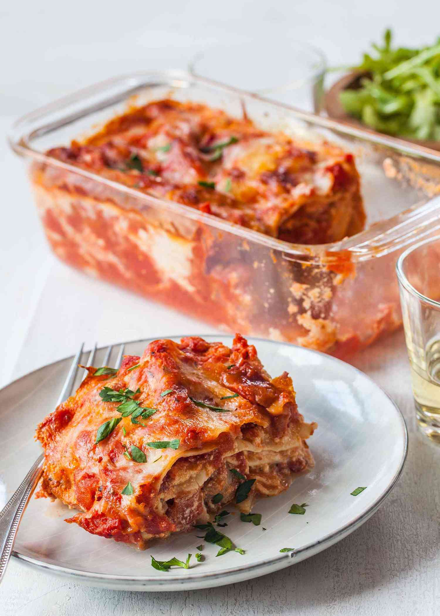 Vertical view of small batch of loaf pan lasagna with sausage on a plate garnished with chopped parsley and a fork on the left. Above the plate is the loaf pan with some of the lasagna missing. A partial view of a bowl of lettuce and a tumbler of white wine is visible.