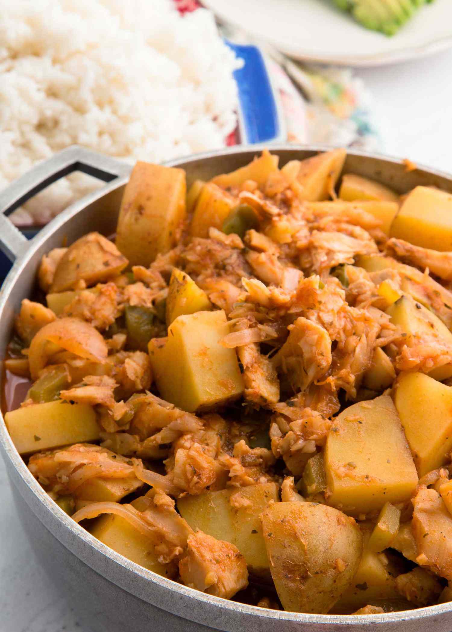 A close up view of a heaping pot of lenten cod. A blue casserole dish with white rice is behind it. A plate with avocado is in the upper right corner.