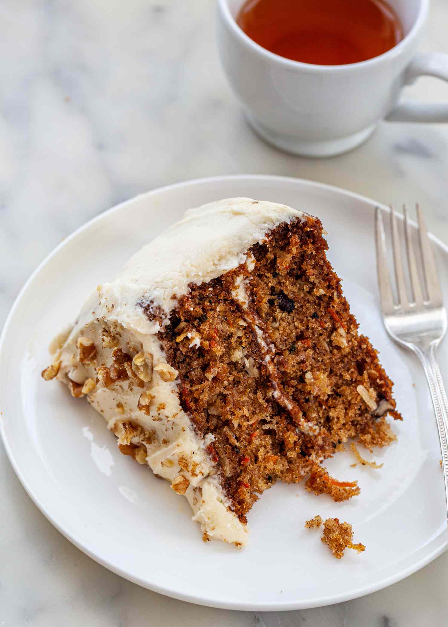 A white plate with a slice of carrot cake on it and a fork, with a cup of tea to the side