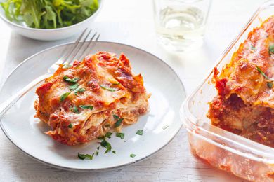 Horizontal view of white plate with slice of small batch lasagna and garnished with chopped parsley. A partial view of the baked lasagna in the glass pan is to the right of the plate. Above the plate to the left is a small bowl of lettuce and a tumbler. A fork is on the left of the plate.