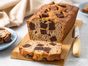 On a Wooden Board, Loaf of Brownie Banana Bread with Slices Cut Off, and Next to It, a Plate with a Slice and a Knife