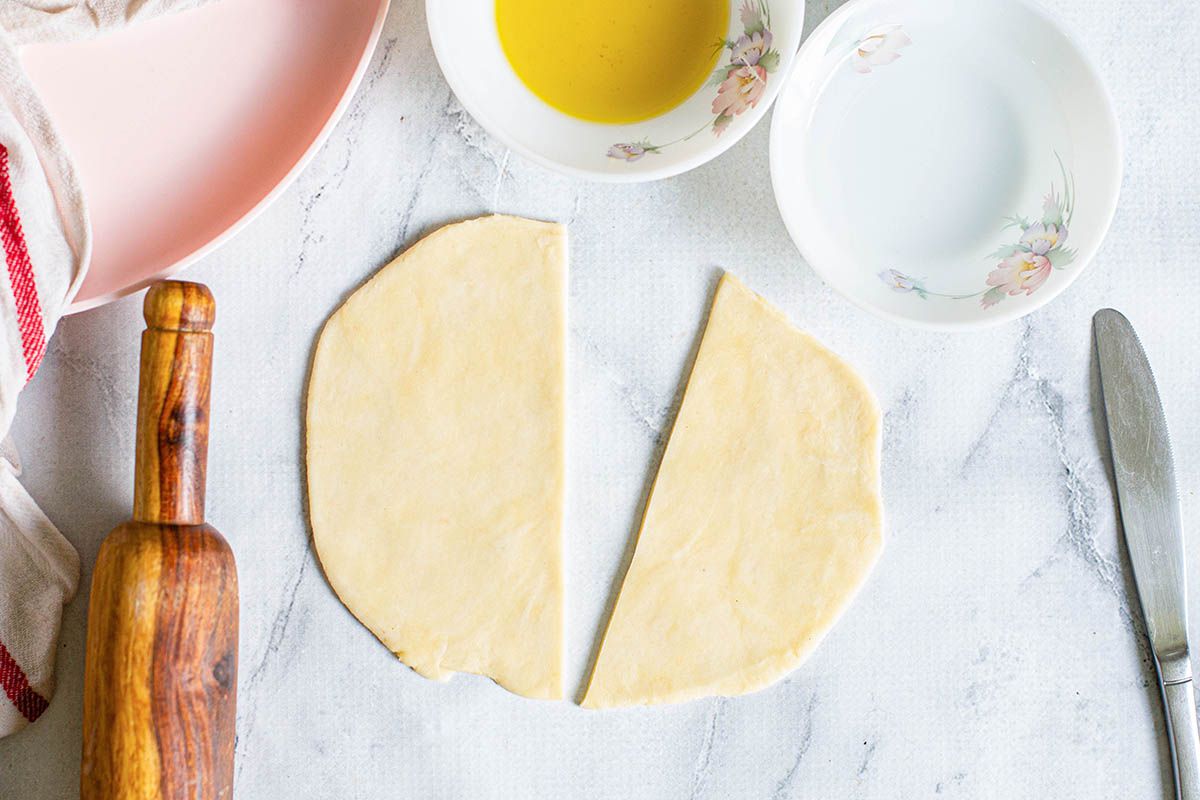 A circle of dough cut in half on a counterop with dishes and a rolling pin nearby.