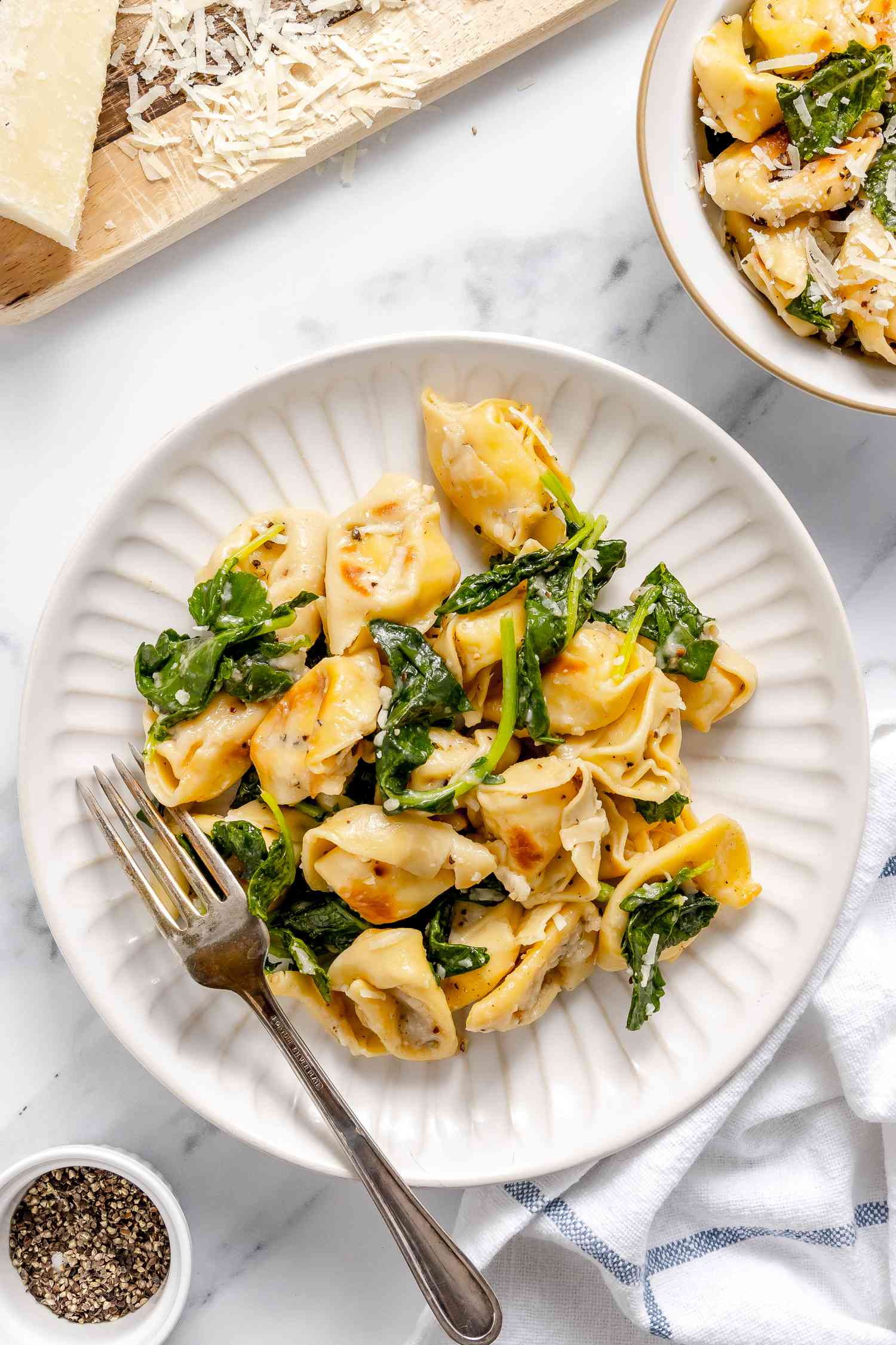 Serving of Skillet Cacio e Pepe Tortellini With Wilted Greens on a Plate With a Fork, and in the Surroundings, a Small Bowl of Cracked Pepper, a Bowl With Another Serving, and a Block of Pecorino Romano on a Cheese Board With Some Freshly Grated Cheese